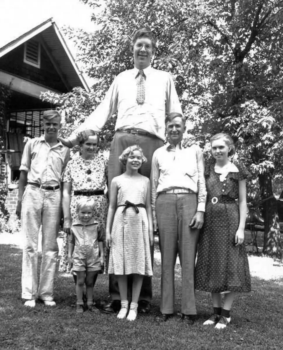 A very tall man stands with his arm resting on an older woman, surrounded by five other people, including children, posing together outside in front of a house and trees on a sunny day.