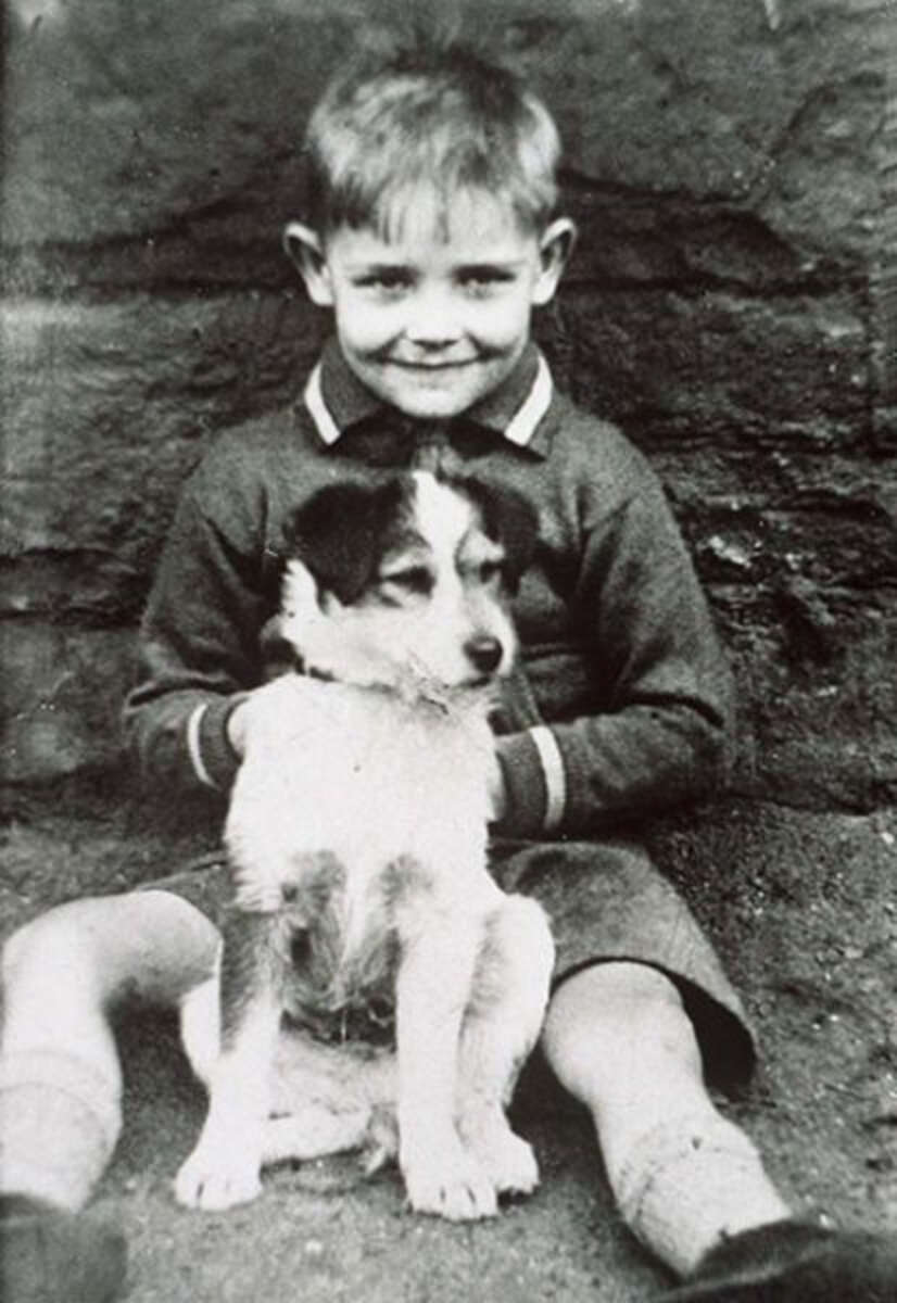A young boy sits on the ground in front of a stone wall, smiling, with his arms around a small dog sitting between his legs. The photo is in black and white.