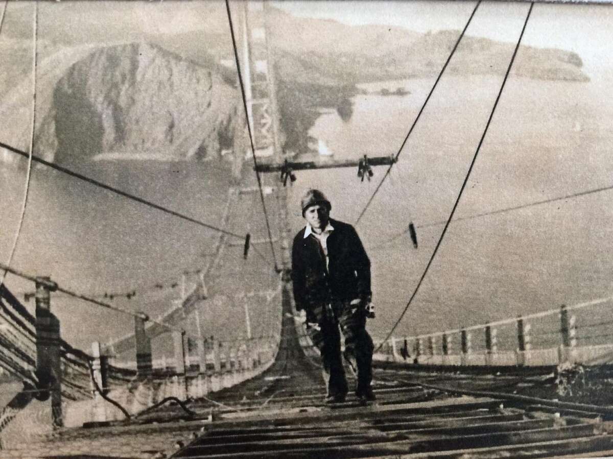 A construction worker stands on the unfinished cables of the Golden Gate Bridge, high above the water, with the bridge’s towers and a rocky shoreline visible in the background. The photo is in black and white.