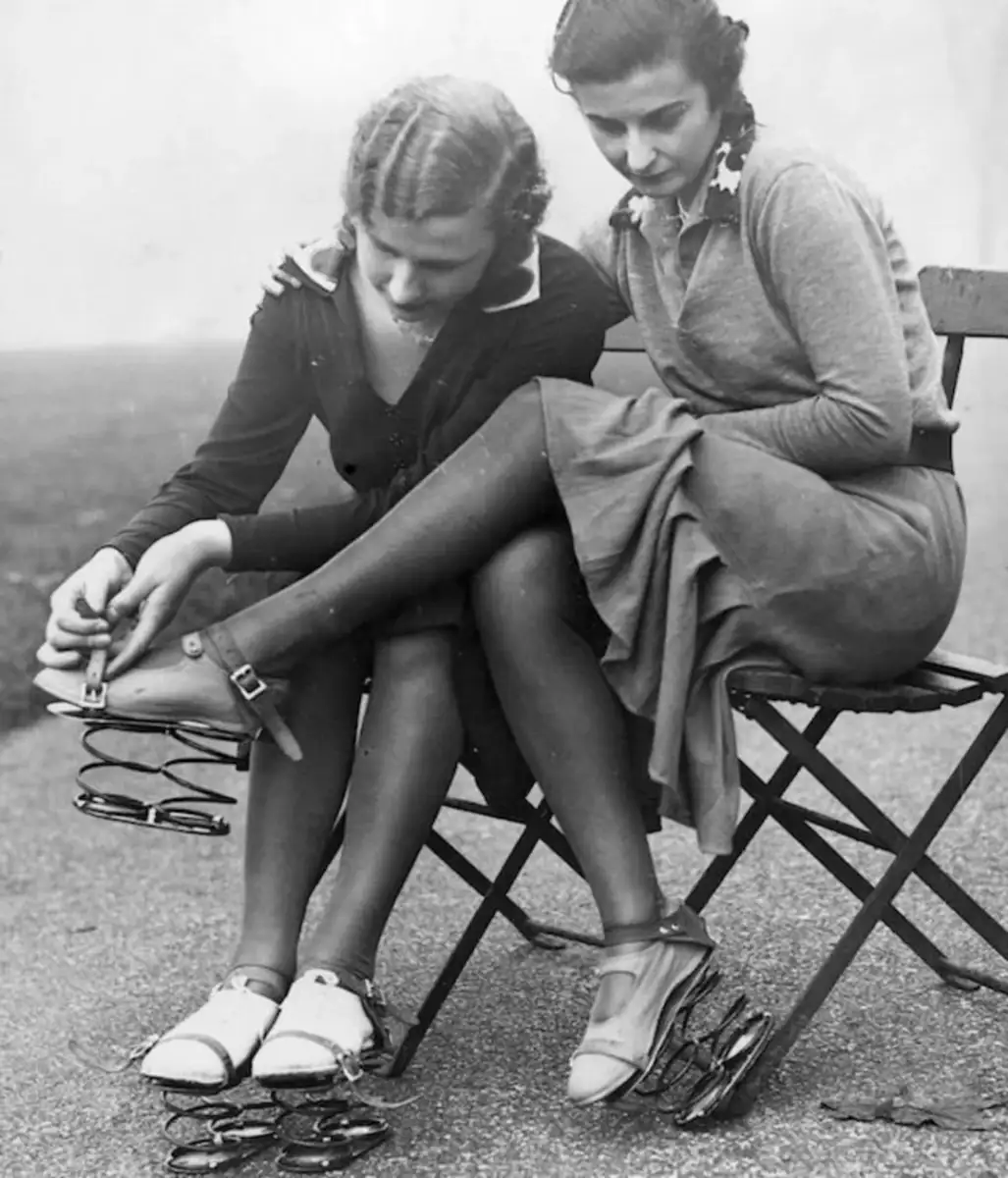 Two women sit on folding chairs outdoors, stacking several pairs of eyeglasses on the foot of one woman. Both are focused on the glasses, with more pairs scattered on the ground near their feet.
