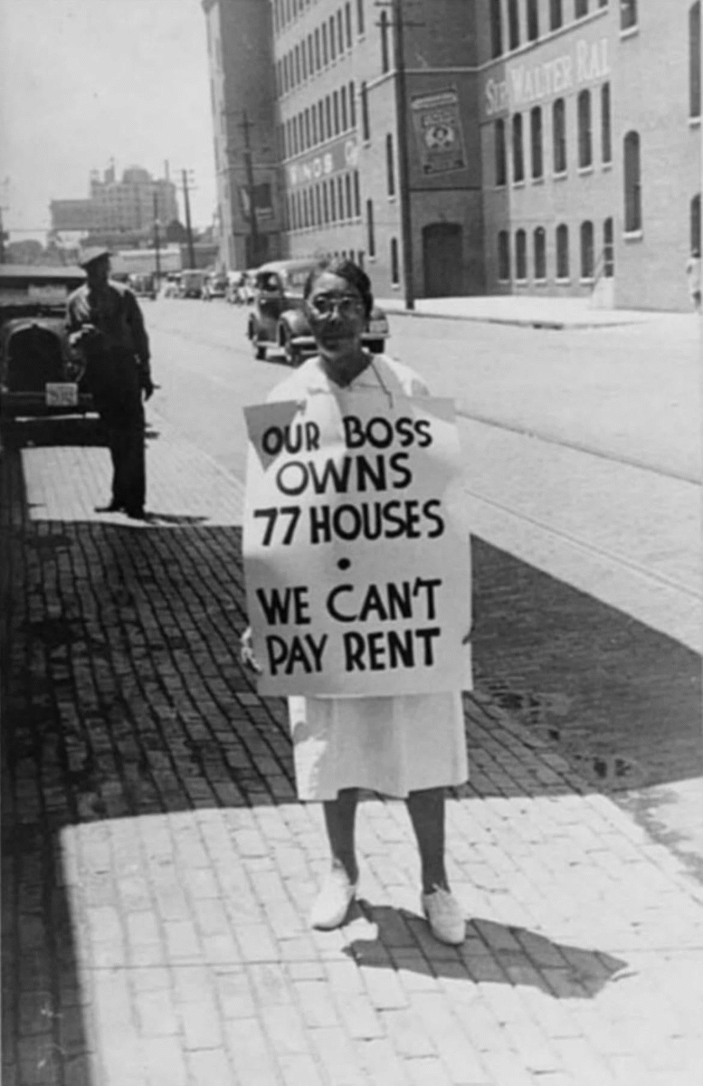 A woman stands on a city sidewalk holding a sign that reads, "Our boss owns 77 houses. We can't pay rent." Old cars and brick buildings are visible in the background.