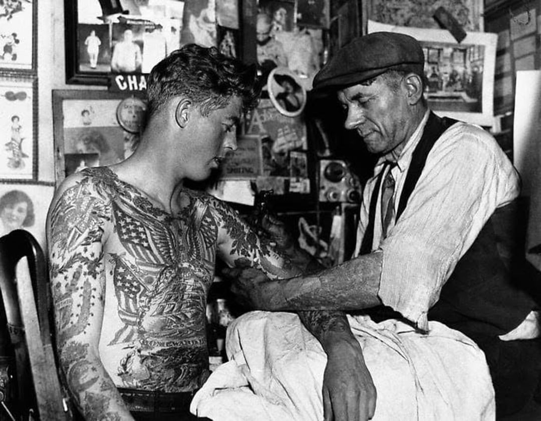 A tattoo artist wearing a cap works on a young man’s arm in a cluttered studio; the man’s chest and arms are heavily tattooed, and walls are covered with photos and artwork. Black and white, vintage setting.