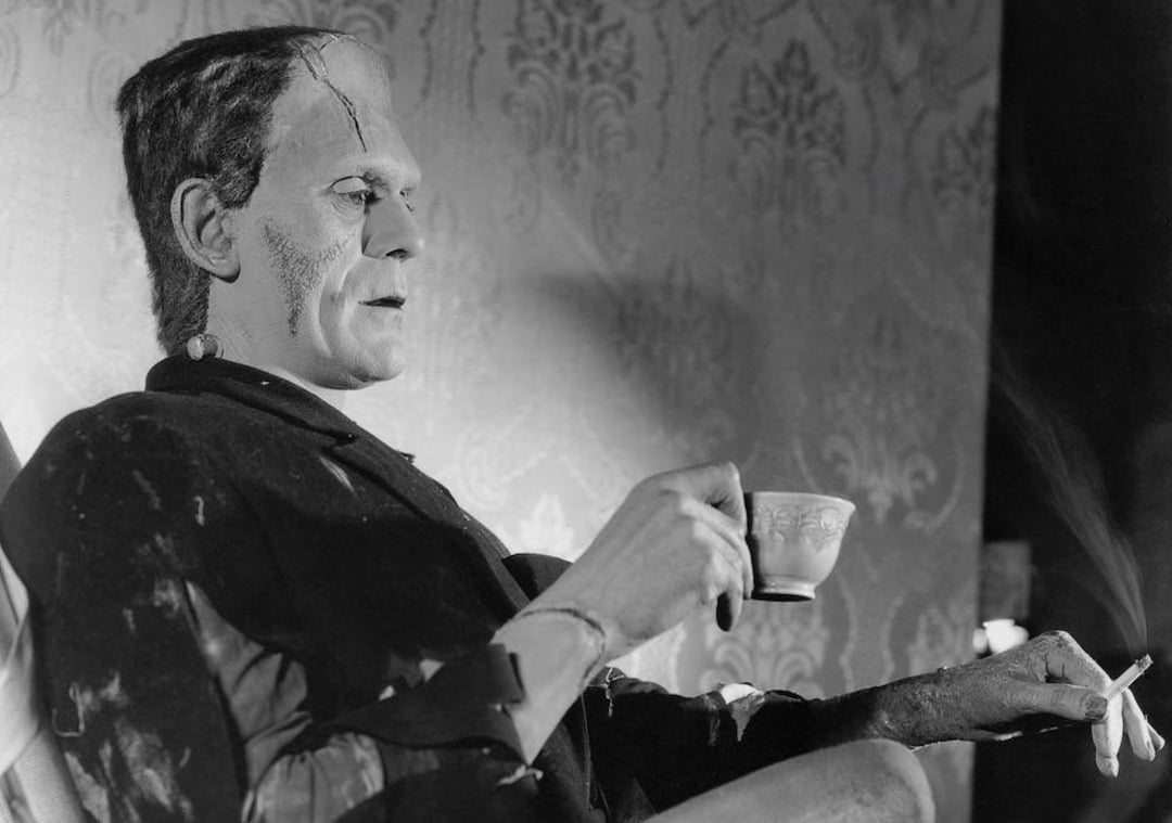 A person in a Frankenstein’s monster costume, complete with scars and bolts, sits against a patterned wall, holding a teacup in one hand and a cigarette in the other, looking relaxed.