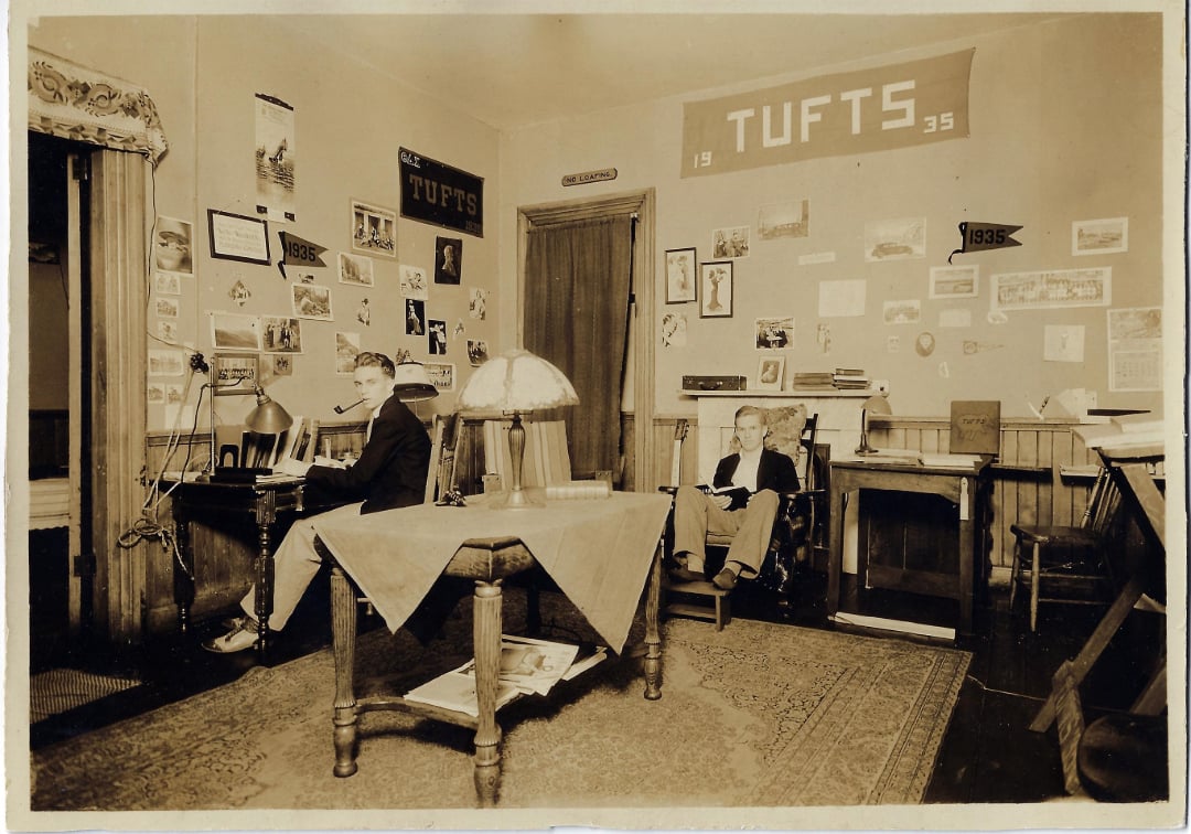 Two men sit in a vintage dorm room decorated with Tufts University banners, photos, and posters on the walls. One man works at a desk, while the other sits in a chair reading. A table with a lamp is in the center.