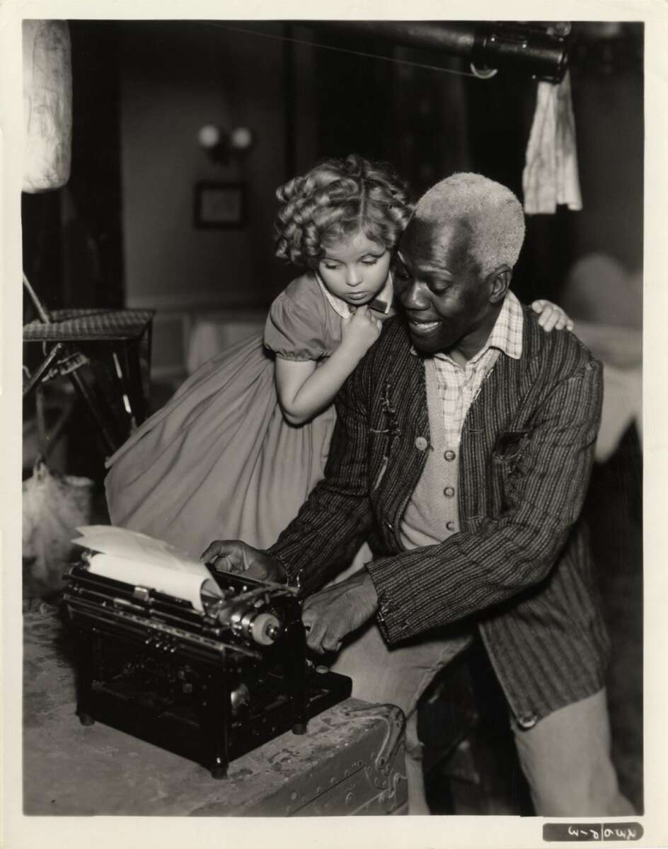 A young girl in a dress stands beside an older man with gray hair who is smiling as he types on a typewriter. The girl looks at the typewriter with interest, resting her hand on the man's shoulder.