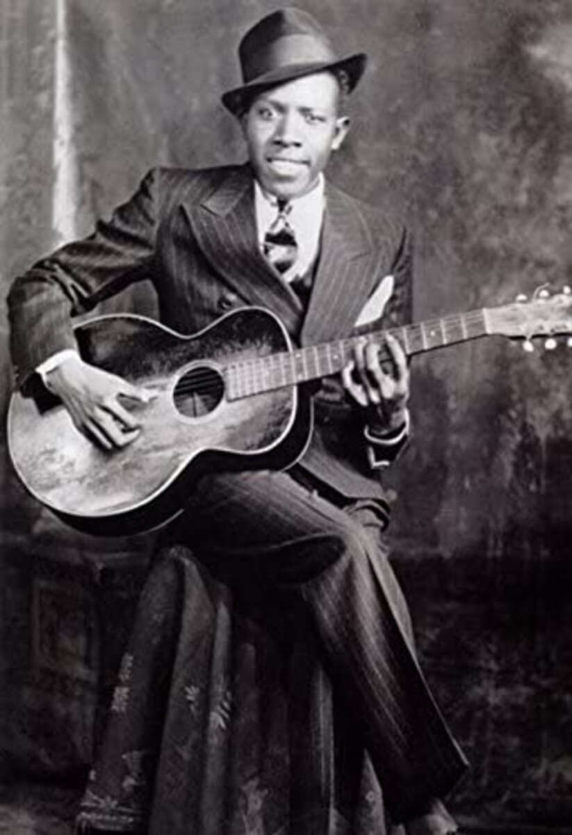 Black-and-white photo of a man in a pinstripe suit and hat, seated and playing an acoustic guitar, looking toward the camera with a serious expression.