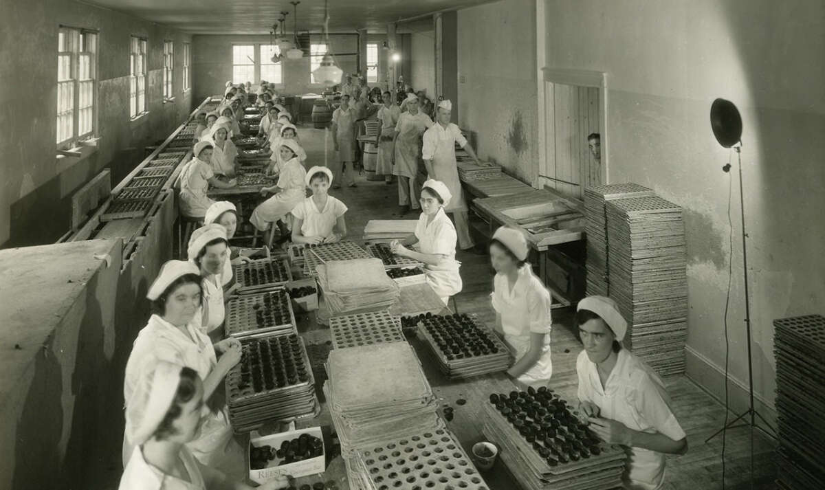 Workers in white uniforms and caps assemble chocolates on trays in a large, crowded factory room with long tables, trays stacked on racks, and sunlight streaming through windows on the left.