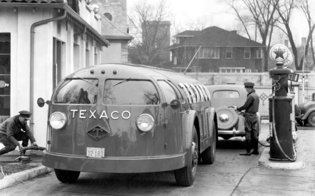 A vintage Texaco bus stops at an old gas station. One attendant checks a tire, while another pumps gas into a nearby car. Leafless trees and houses are seen in the background. The scene appears to be from the early 20th century.