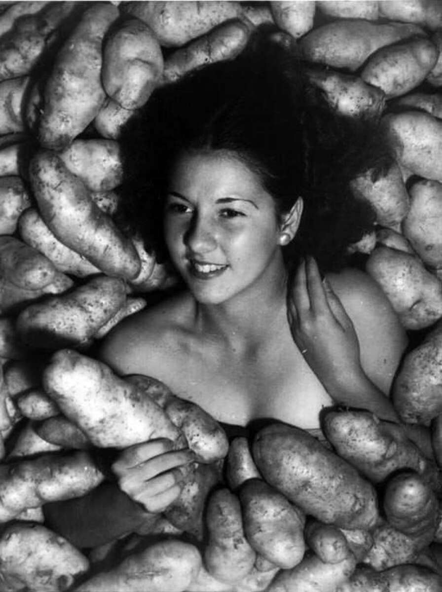 A young woman smiles while lying among a large pile of potatoes, with her head and shoulders visible and her hand holding one of the potatoes.