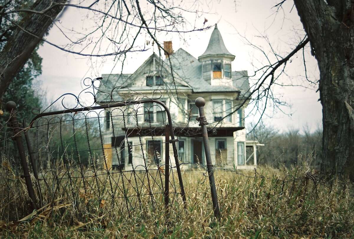An old, abandoned Victorian-style house with boarded windows stands behind a broken, rusted fence, surrounded by tall, overgrown grass and bare trees.