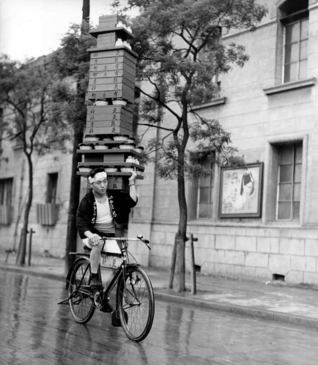 A man rides a bicycle on a wet street, balancing a tall stack of boxes on one hand. He wears traditional clothing and a headband, and the background shows a stone building with trees and a poster.