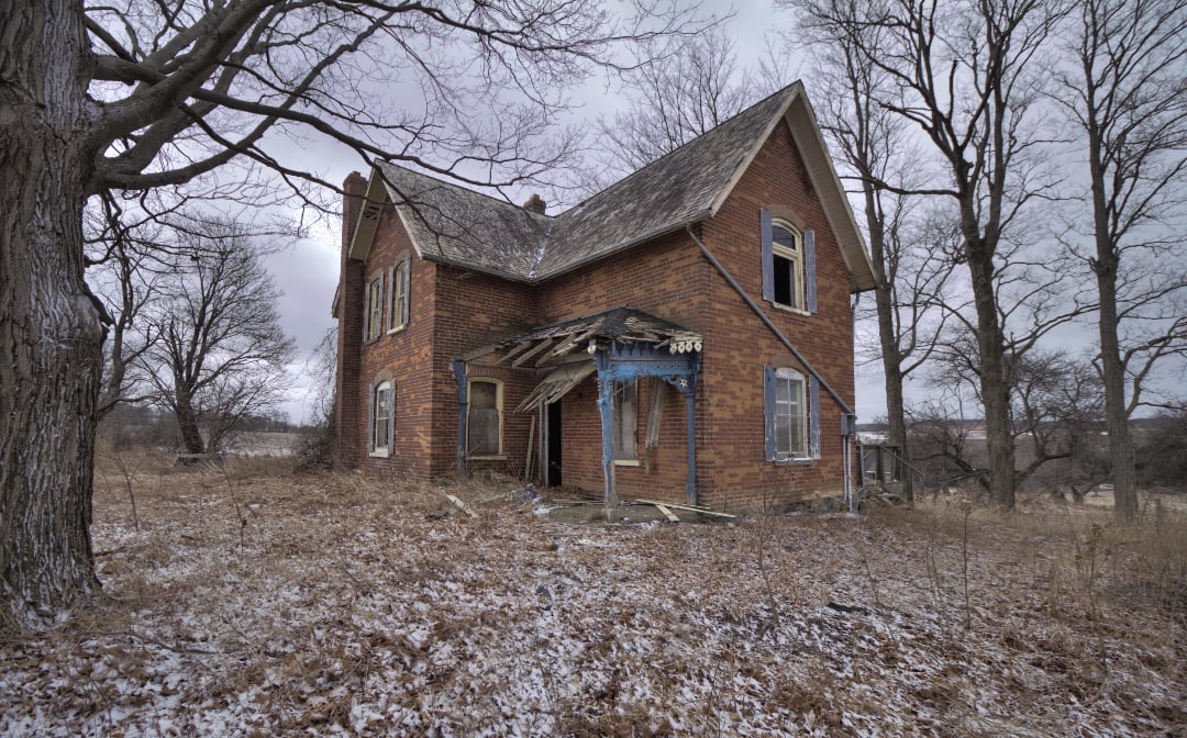 An old, abandoned brick house with broken windows and a collapsing porch stands in a barren, leafless landscape dusted with snow under a cloudy sky.