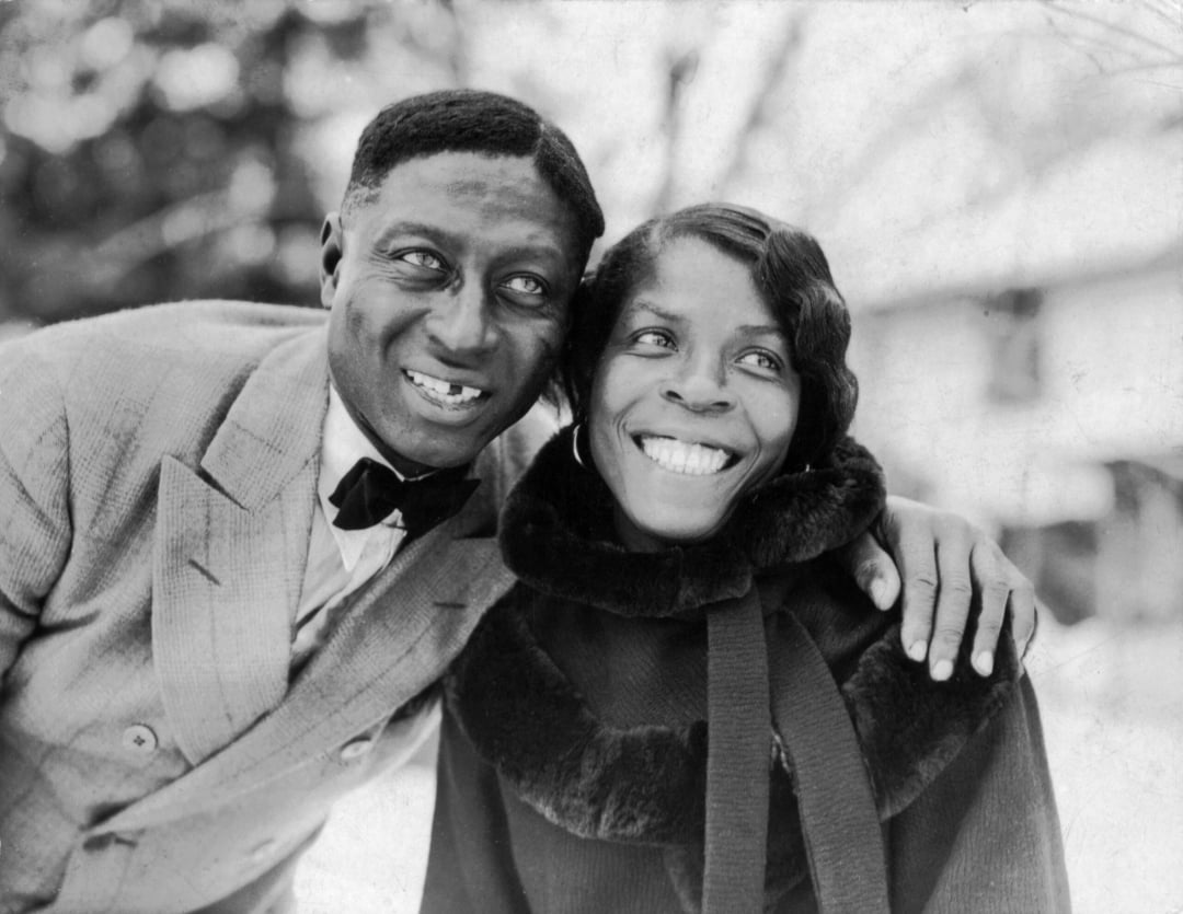 A smiling man in a suit and bow tie stands with his arm around a smiling woman in a fur-collared coat. They are outdoors, and it appears to be winter. Both look happy, posing closely together.