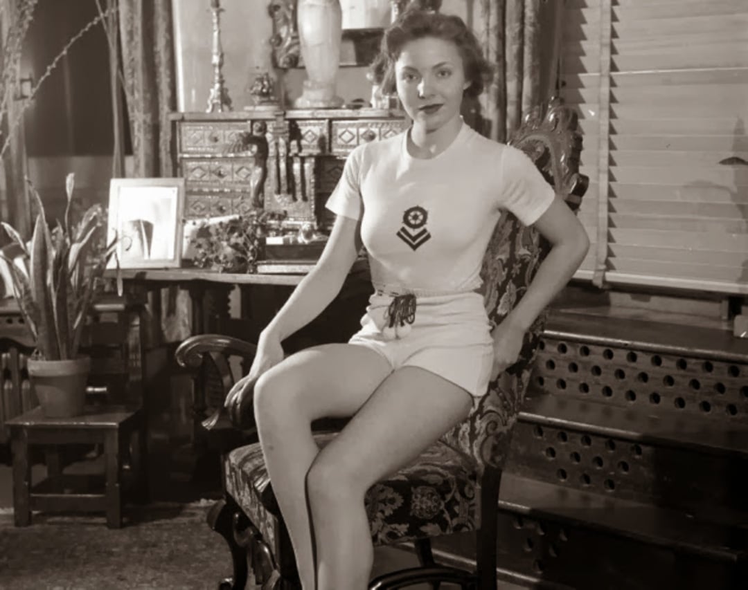 A woman in vintage athletic attire sits on an ornate chair in a well-decorated room with plants, framed photos, and antique furniture. She wears a white shirt and shorts, looking calmly at the camera.