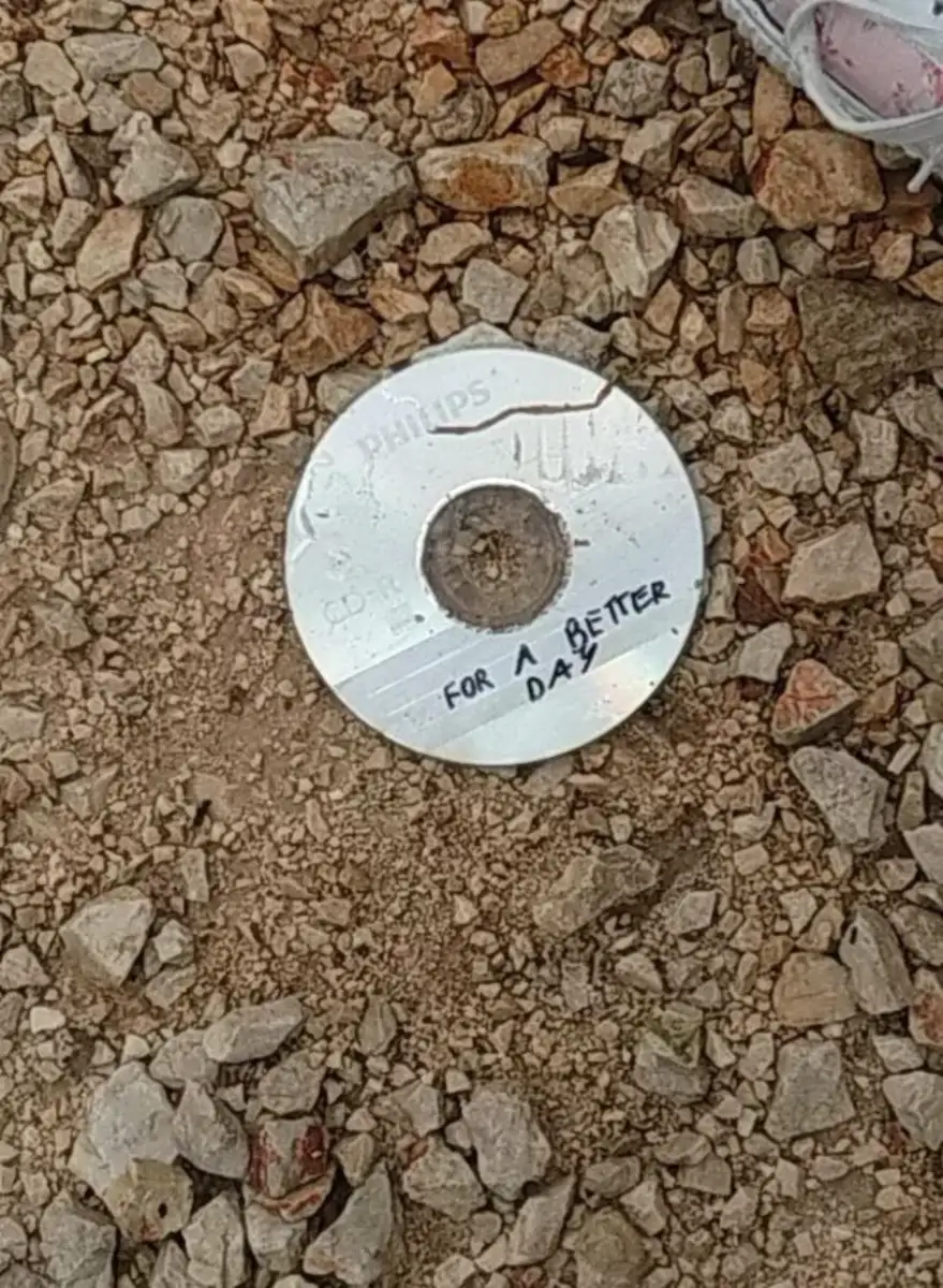 A CD labeled "FOR A BETTER DAY" lies on a gravel surface among small rocks, with part of a sneaker visible at the top right corner of the image.