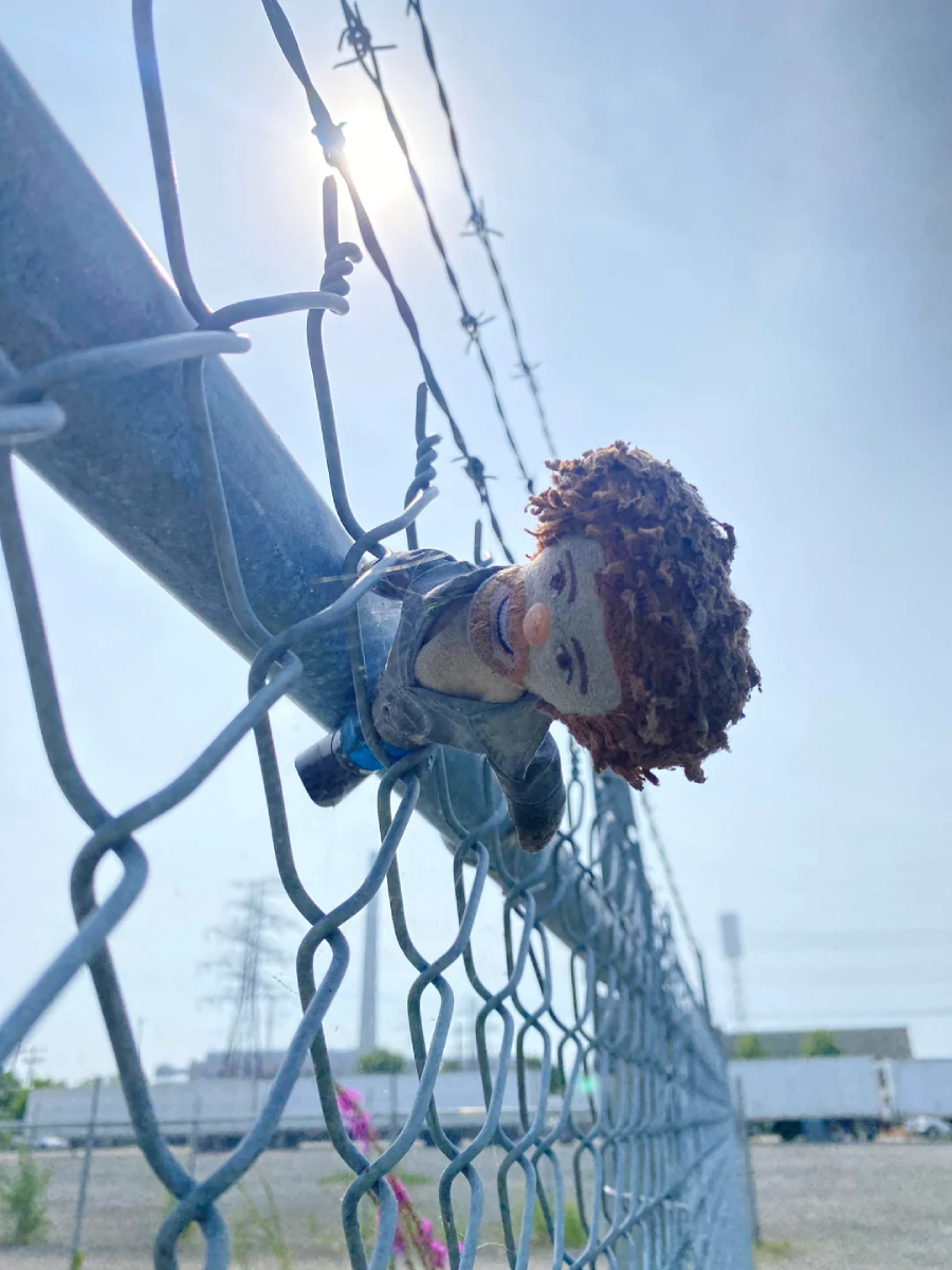A plush doll with curly hair is stuck through the top of a chain-link fence under barbed wire, with sunlight shining in the sky and industrial buildings visible in the background.