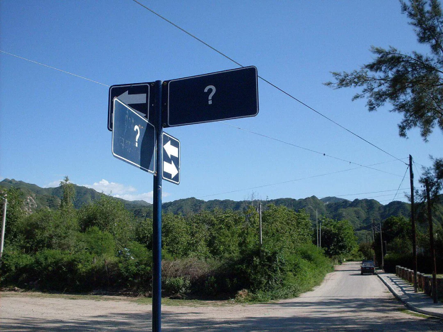 A street sign at a rural intersection with all arrows and panels replaced by question marks, set against a backdrop of green hills and a clear blue sky.