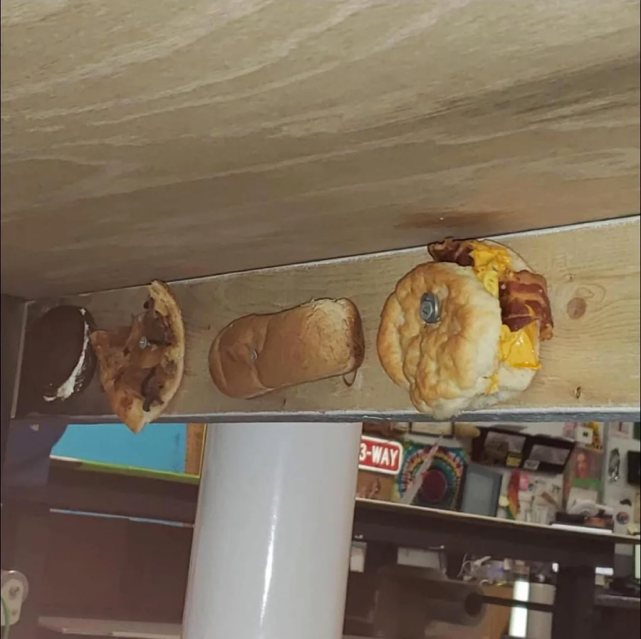 A hamburger, hotdog, Oreo, and slice of pizza are screwed upside down to the underside of a wooden table. The background shows a cluttered room with shelves and signs.
