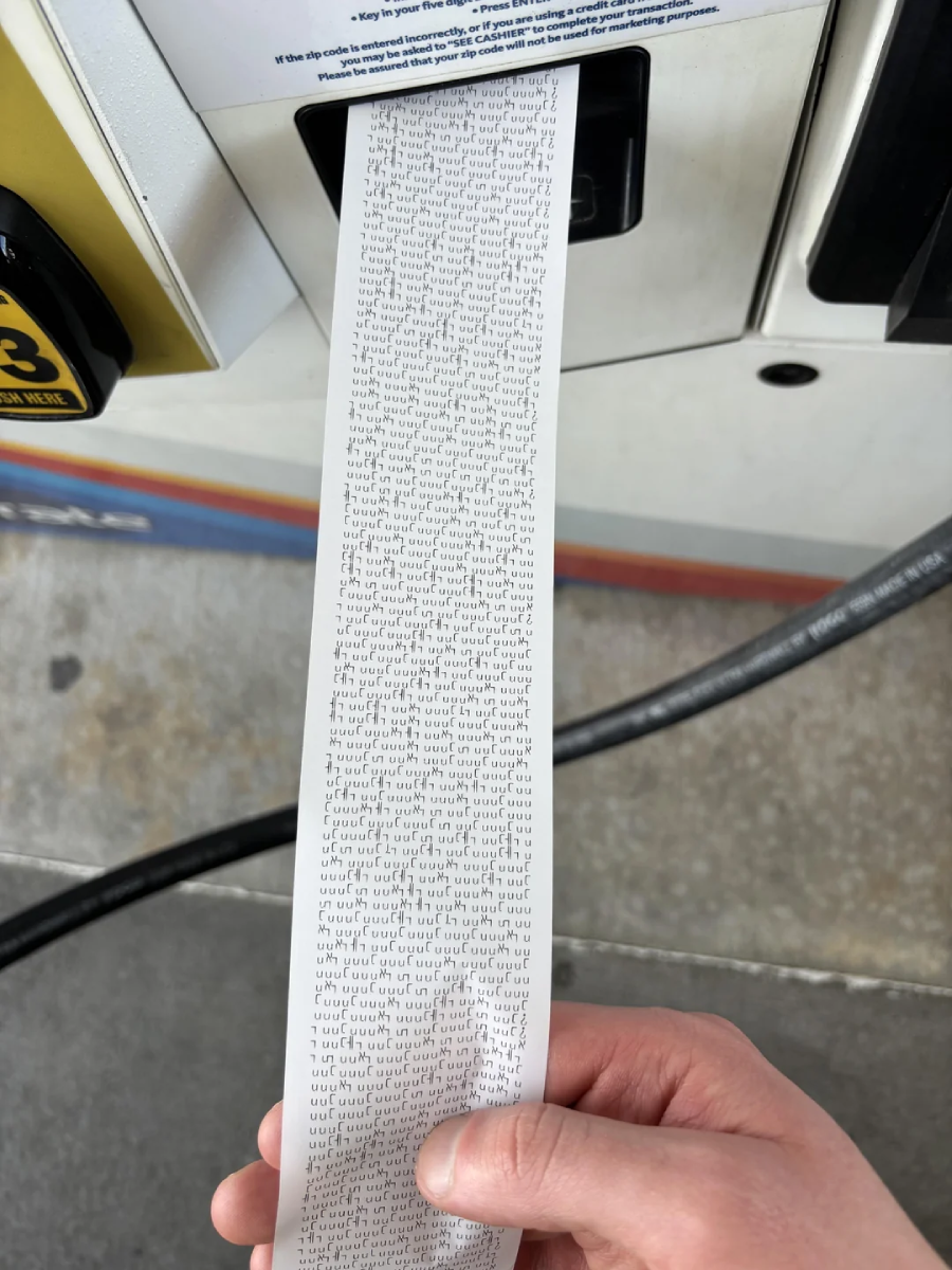 A hand holds a very long gas pump receipt printed with an unusually large amount of small text, while standing at a fuel station pump. The receipt extends well past the dispenser.