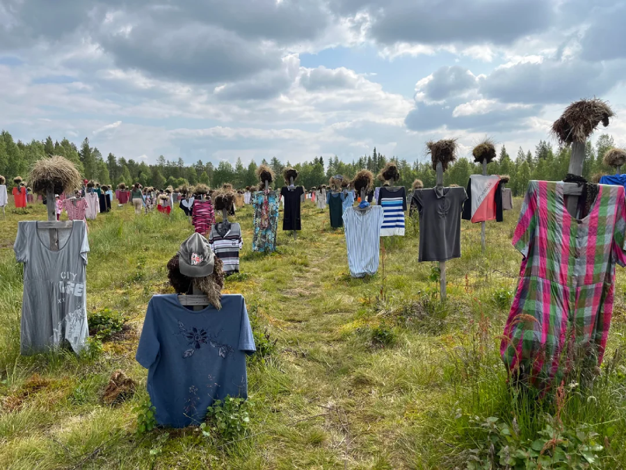 Rows of wooden posts dressed in various shirts and dresses stand in a grassy field under a partly cloudy sky, with hay bundles placed on top of many posts like heads. Trees line the horizon in the background.
