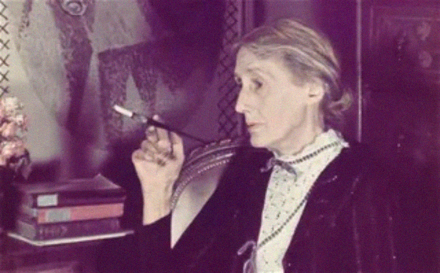 An older woman with light hair sits in profile, holding a cigarette holder. She wears a dark jacket over a light blouse. Books and a framed artwork are visible in the softly lit background.