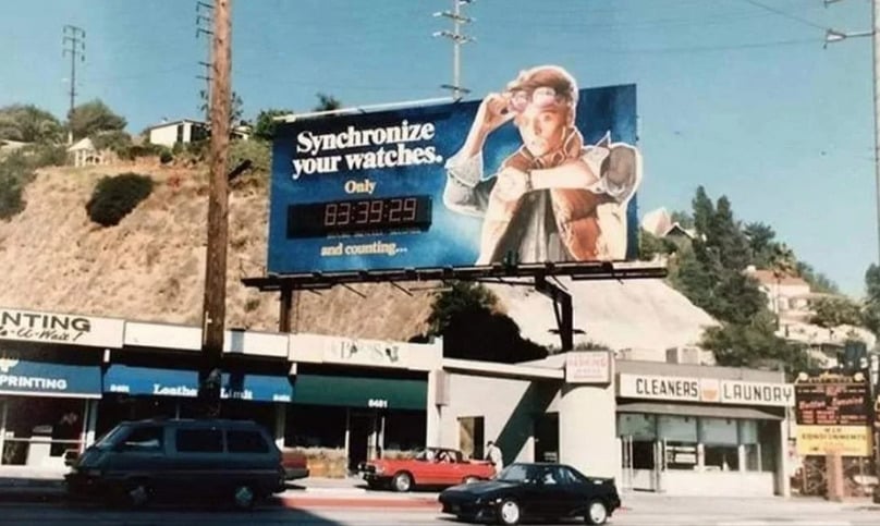 A billboard above a street shows a character in a red vest and jeans adjusting his watch with the text “Synchronize your watches” and a digital countdown timer. Shops and cars are visible below the sign.
