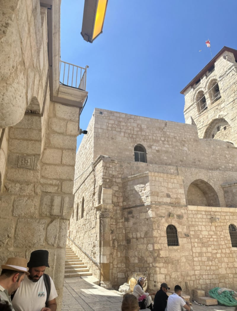 People sit and stand near a historic stone building with arched windows and a tower under a clear blue sky; stone steps lead up the side, and sunlight casts shadows across the scene.
