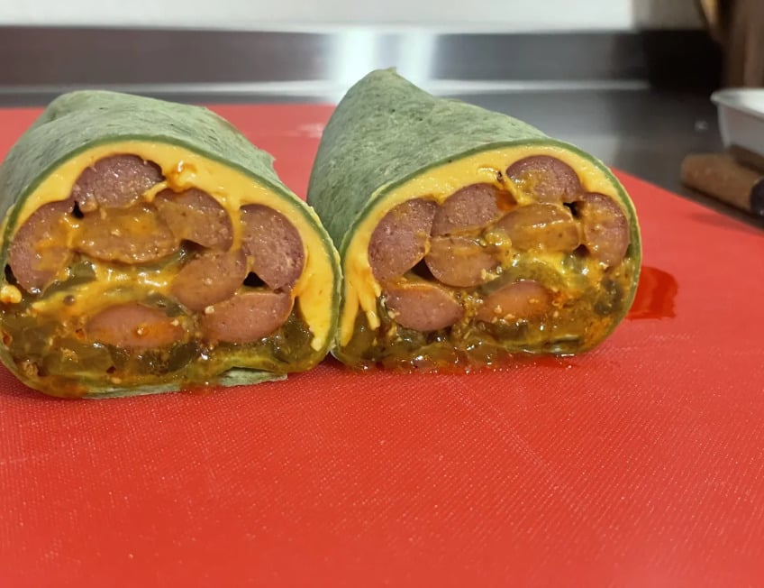 A spinach wrap cut in half, filled with sliced sausage and melted cheese, placed on a red cutting board.