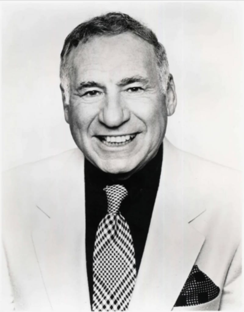 A smiling older man in a light-colored suit, dark shirt, and patterned tie, posing for a black-and-white portrait against a plain background.