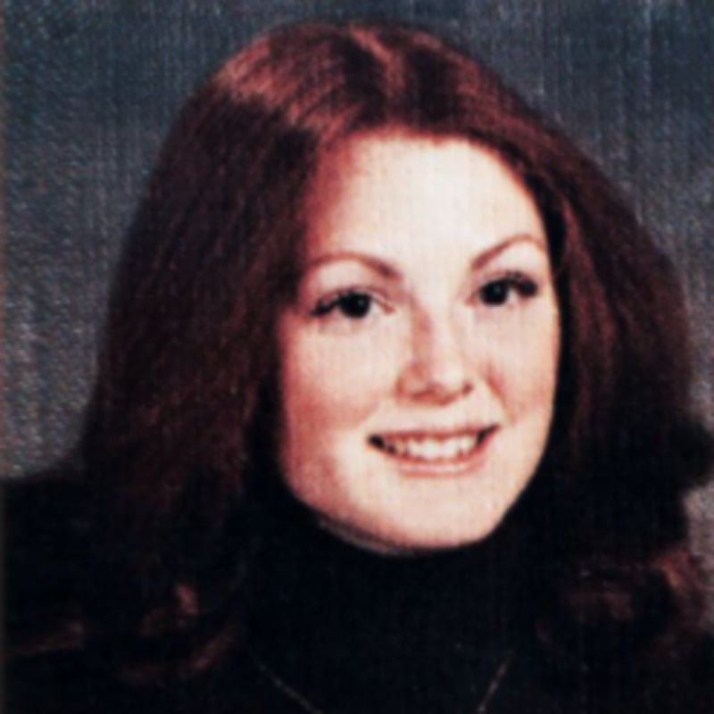 A young woman with long, wavy red hair smiles at the camera. She is wearing a dark top and appears to be posing for a formal portrait, possibly a school or yearbook photo. The background is gray and softly textured.