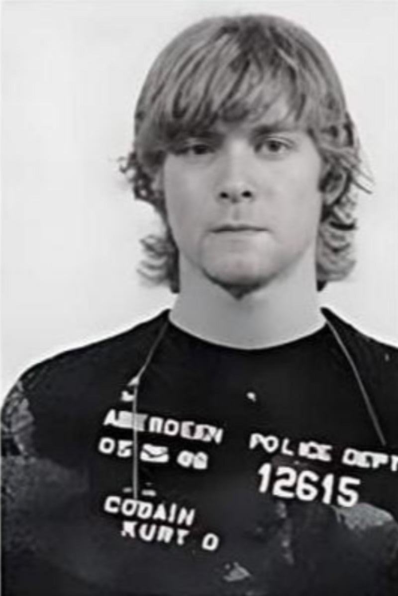 Black and white photo of a young man with wavy, shoulder-length hair, facing the camera with a neutral expression. He is wearing a shirt with police department text and numbers, resembling a mugshot.
