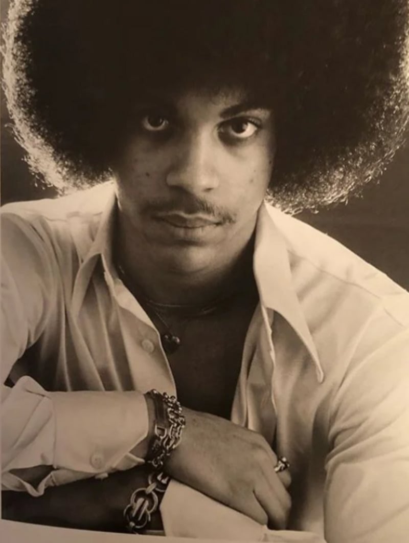 Black and white photo of a young man with an afro hairstyle, wearing an open-collared shirt and several bracelets, looking directly at the camera with a serious expression.