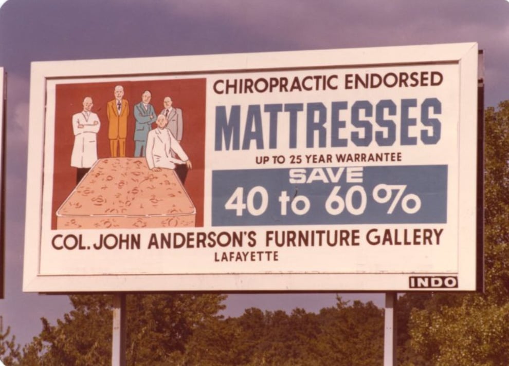 A billboard advertises mattresses with 40-60% savings, endorsed by chiropractors, featuring illustrated doctors and a mattress, for Col. John Anderson’s Furniture Gallery in Lafayette.