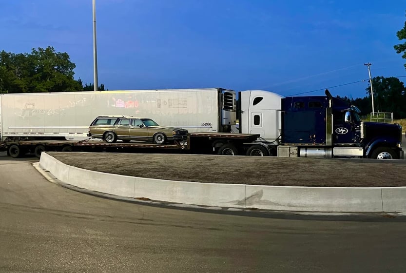 A large semi truck with a white trailer is parked, carrying an old beige station wagon on a flatbed section behind the cab at dusk, next to a curved concrete barrier and trees in the background.