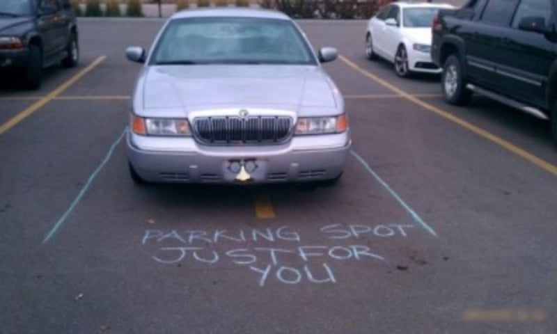 A silver sedan is parked over two parking spaces, with blue chalk lines and the words "PARKING SPOT JUST FOR YOU" written on the pavement in front of the car.