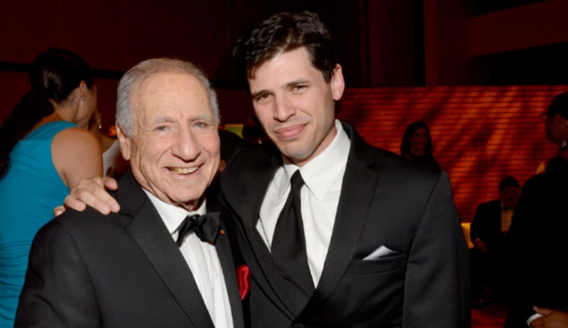 Two men in tuxedos smile and pose together at an indoor formal event. One has his arm around the other’s shoulder. Blurred people and warm lighting are visible in the background.