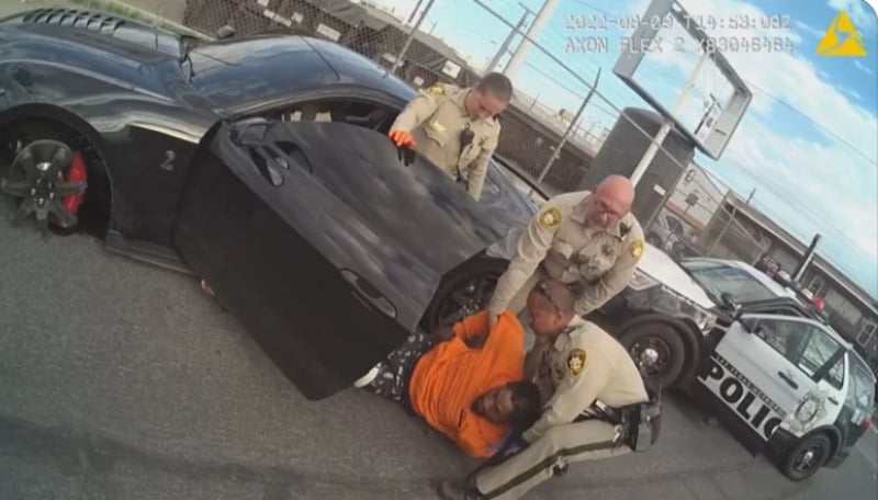 Three police officers detain a man in an orange shirt on the ground beside a black car with an open door. A police vehicle is parked nearby in a fenced area.