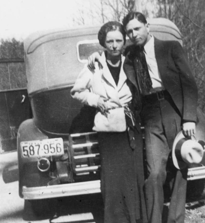 A black-and-white photo of a man and woman standing close together in front of an old car. The man has his arm around the woman and is holding a hat. Both wear 1930s-style clothing and look directly at the camera.