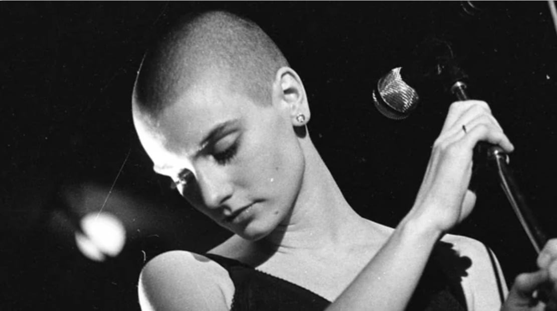 A person with a shaved head and earrings stands in front of a microphone, looking downward with eyes closed, holding the mic stand with one hand in a dimly lit setting.