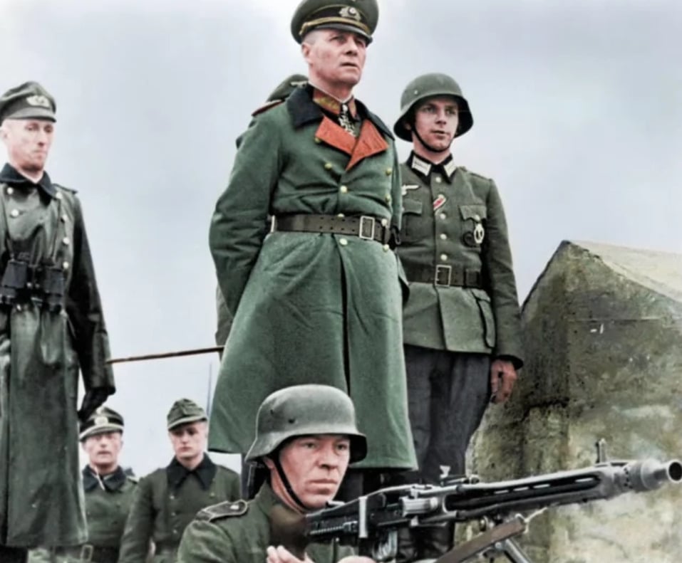 A group of German soldiers in World War II uniforms, including one high-ranking officer in the center, stands outdoors. One soldier in the foreground operates a machine gun, while others stand at attention behind him.