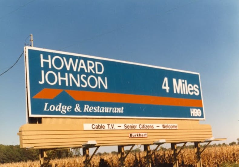 A large roadside billboard reads "Howard Johnson Lodge & Restaurant 4 Miles" with an HBO logo. Underneath, a sign says "Cable T.V. – Senior Citizens – Welcome". The billboard stands by a cornfield under a clear sky.