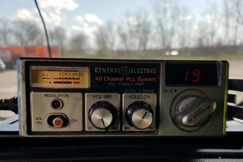 A close-up of a vintage General Electric 40 Channel PLL System radio with dials for volume, squelch, and modulation. The digital display shows the number 19, and trees are visible through the window behind it.