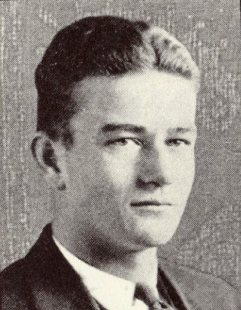 Black and white portrait of a young man with short, slicked-back hair, wearing a suit and tie, looking slightly to the side with a neutral expression.