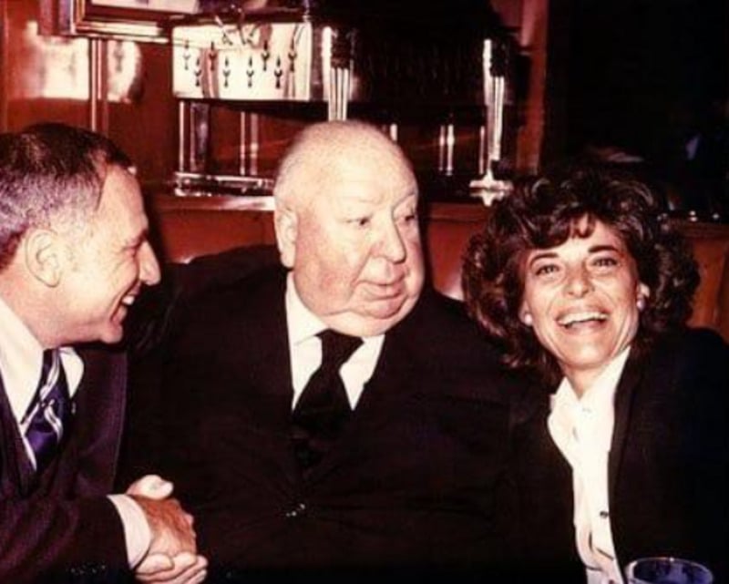 Three people are sitting close together, smiling and laughing. The man in the center, older and bald, looks serious while the man on the left and the woman on the right are both smiling happily in a warmly lit setting.