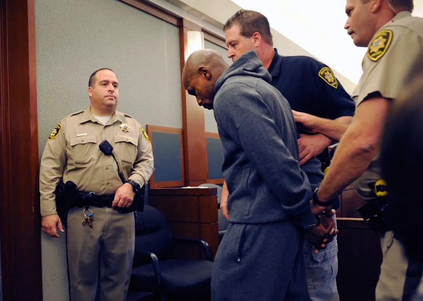 Three uniformed officers escort a handcuffed man in a gray tracksuit inside a courtroom. The man looks down while one officer holds his arm and another stands by the door, watching.
