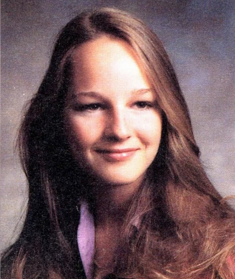 A young woman with long, light brown hair smiles softly, wearing a light-colored collared blouse. The background is a softly blurred gradient of blue and brown, resembling a classic school portrait.