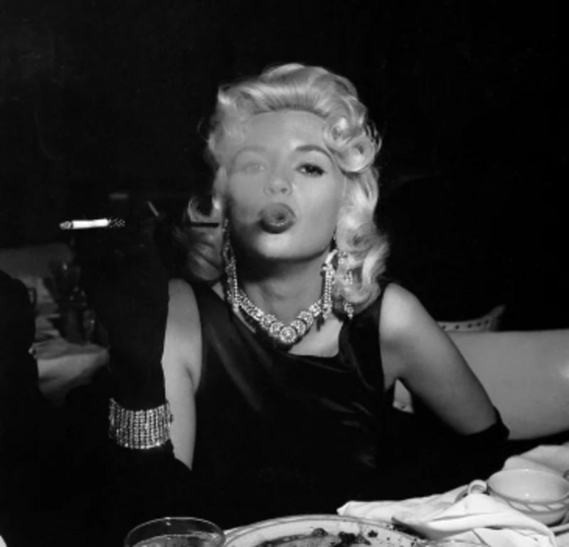 A glamorous woman with blonde hair in waves, wearing a dark dress, sparkling jewelry, and gloves, sits at a table and blows smoke from a cigarette, with a confident expression.