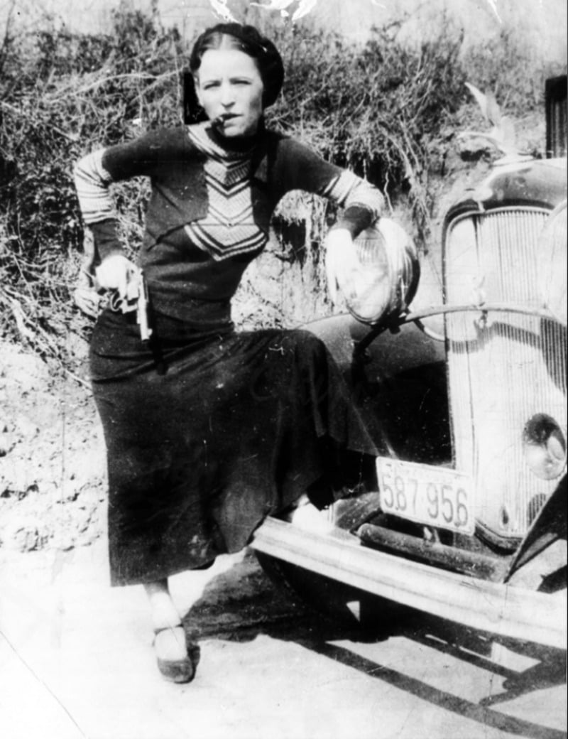 A woman in a long dress stands with one foot on the bumper of an old car, holding a gun and looking at the camera. The scene appears vintage, with bushes and dirt in the background.