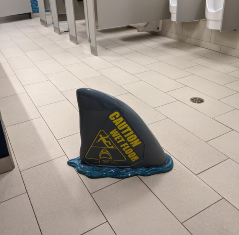 A restroom with a "Caution Wet Floor" sign shaped like a shark fin surrounded by blue puddle-like edges, placed on beige tiled flooring near stalls and urinals.