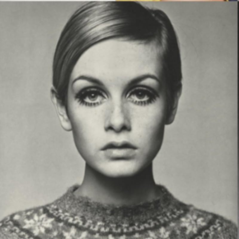 Black and white portrait of a young woman with short, neatly parted hair. She wears a patterned sweater and has dramatic eyeliner accentuating her eyes as she looks directly at the camera with a neutral expression.